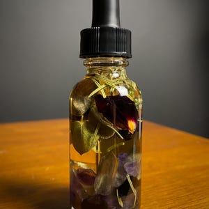 May include: A clear glass bottle with a black dropper top, filled with a golden liquid and botanical elements. The bottle contains leaves, purple crystals, and a dark red flower. The bottle sits on a wooden surface.