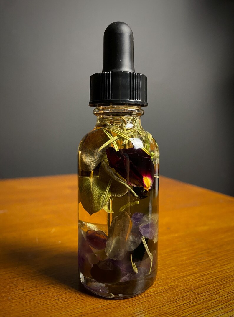 May include: A clear glass bottle with a black dropper top, filled with a golden liquid and botanical elements. The bottle contains leaves, purple crystals, and a dark red flower. The bottle sits on a wooden surface.
