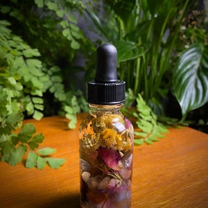 May include: A clear glass bottle with a black dropper top, filled with a golden-hued liquid and dried flowers. The bottle contains pink rose petals, yellow flowers, and other botanicals. The background features green foliage.