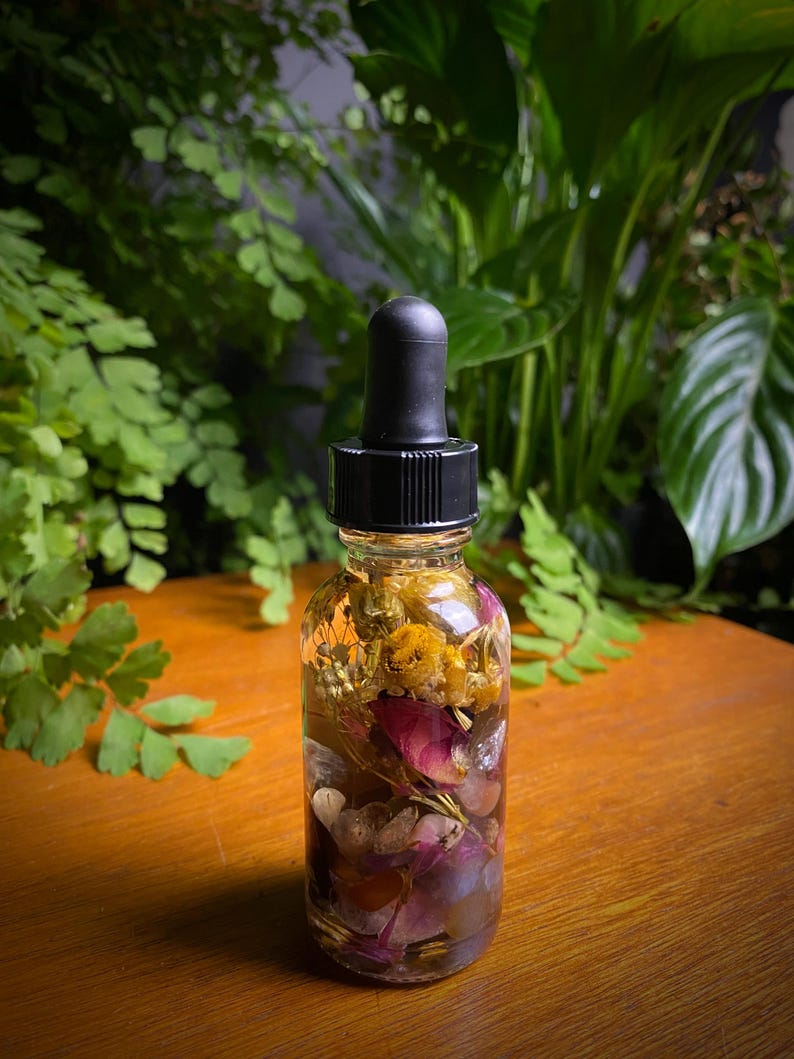May include: A clear glass bottle with a black dropper top, filled with a golden-hued liquid and dried flowers. The bottle contains pink rose petals, yellow flowers, and other botanicals. The background features green foliage.