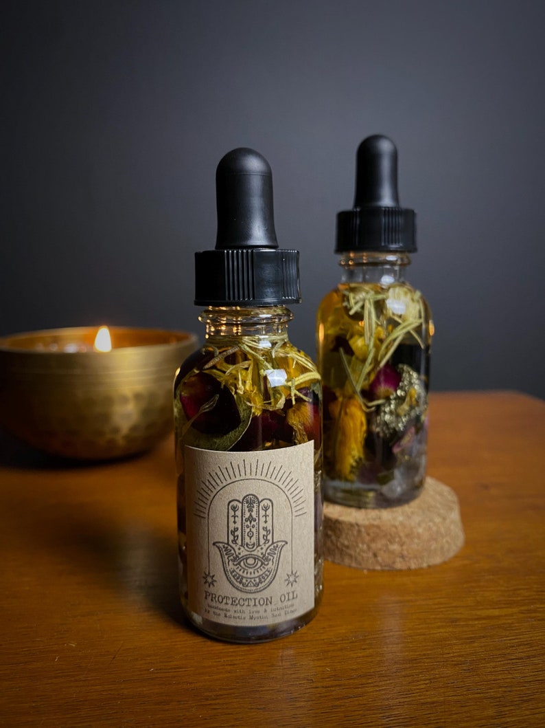 May include: Two small glass bottles filled with dried flowers and herbs. The bottles have black dropper tops and are labeled "Protection Oil" with a hamsa hand symbol. The bottles are sitting on a wooden surface.
