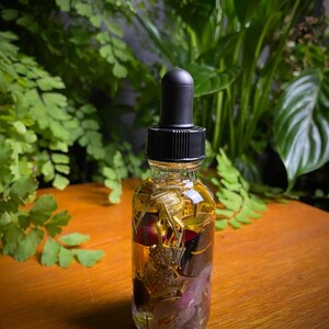 May include: A clear glass bottle with a black dropper top filled with a golden liquid and various dried botanicals, including flowers, leaves, and herbs.