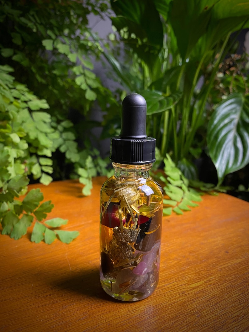 May include: A clear glass bottle with a black dropper top filled with a golden liquid and various dried botanicals, including flowers, leaves, and herbs.