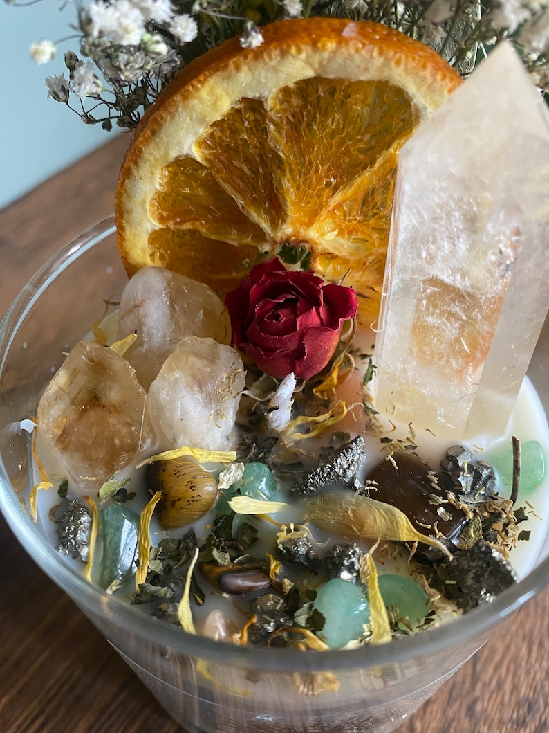 May include: A clear glass jar filled with a white candle, dried orange slices, a red rose, and various crystals and herbs. The jar is sitting on a wooden surface.