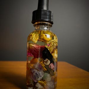May include: A clear glass bottle filled with a yellow liquid, dried flowers, and colorful stones. The bottle has a black dropper top. The contents are visible through the glass, creating a layered effect. The bottle sits on a wooden surface.