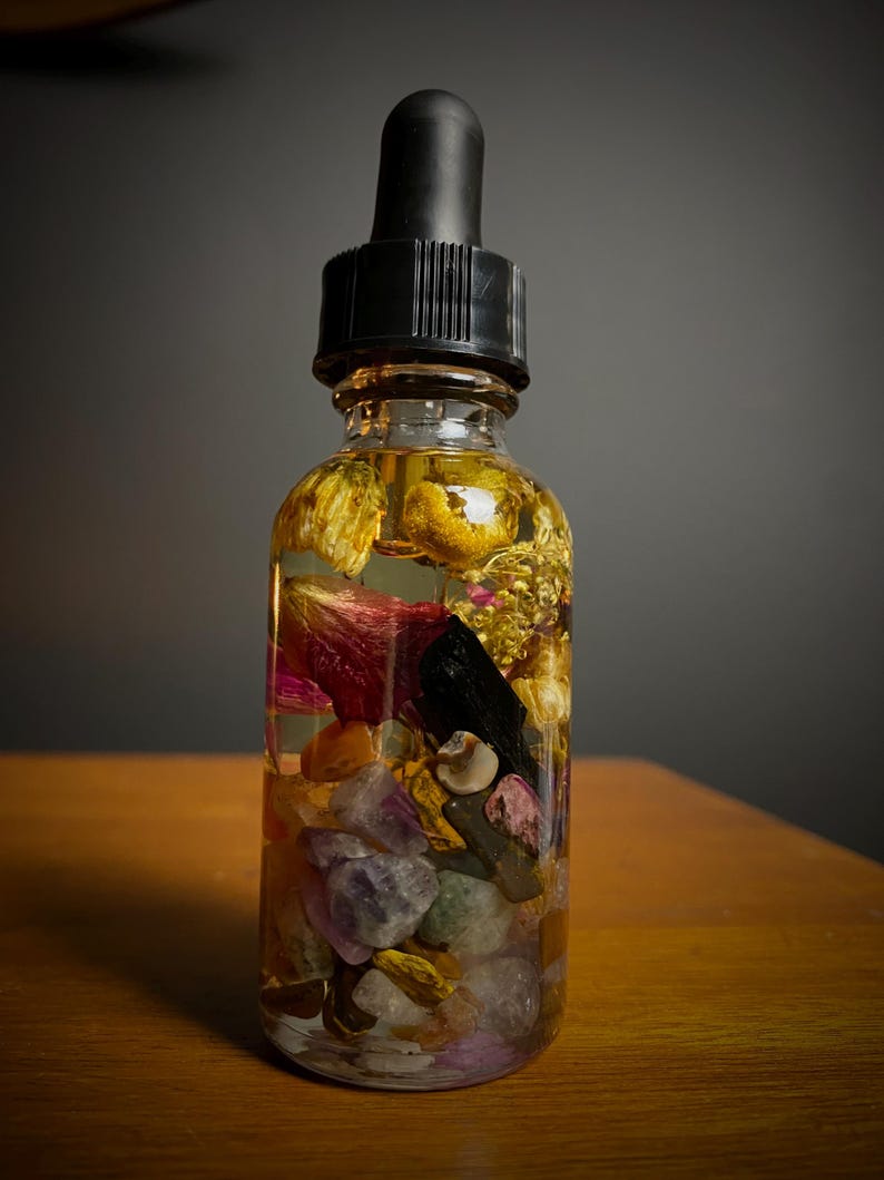 May include: A clear glass bottle filled with a yellow liquid, dried flowers, and colorful stones. The bottle has a black dropper top. The contents are visible through the glass, creating a layered effect. The bottle sits on a wooden surface.