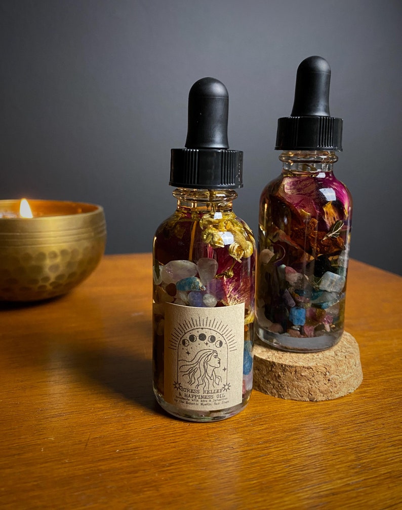 May include: Two small glass bottles filled with dried flowers, crystals, and a clear liquid. The bottles have black dropper tops and are labeled "Stress Relief & Happiness Oil" with a moon and star design. The bottles are sitting on a cork coaster.