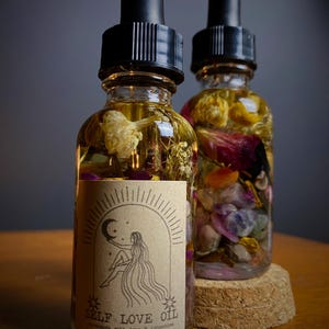 May include: Two glass bottles of oil with black droppers. The bottles are filled with flowers, crystals, and herbs. One bottle has a label that reads "SELF LOVE OIL" with an illustration of a woman. The bottles sit on a wooden surface.