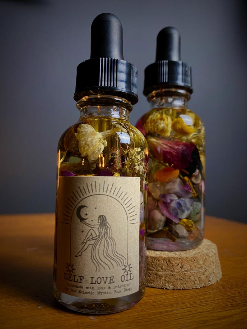 May include: Two glass bottles of oil with black droppers. The bottles are filled with flowers, crystals, and herbs. One bottle has a label that reads "SELF LOVE OIL" with an illustration of a woman. The bottles sit on a wooden surface.