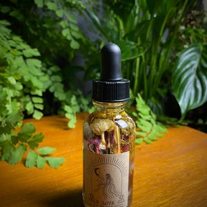 May include: A clear glass bottle with a black dropper top filled with a light yellow oil and dried flowers. The bottle has a brown paper label with the text "SELF LOVE OIL" and a drawing of a woman with long hair sitting on a crescent moon.