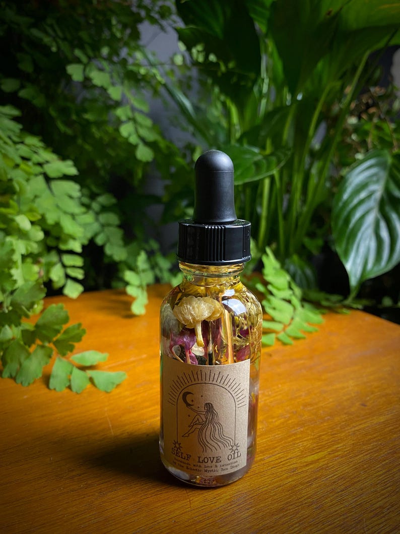 May include: A clear glass bottle with a black dropper top filled with a light yellow oil and dried flowers. The bottle has a brown paper label with the text "SELF LOVE OIL" and a drawing of a woman with long hair sitting on a crescent moon.