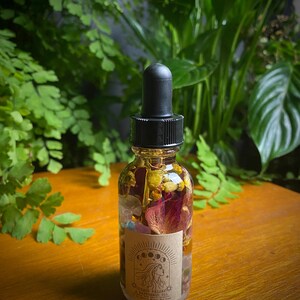 May include: A clear glass bottle with a black dropper top filled with a golden liquid and dried flowers and crystals. The label reads "WITCHY DAILY HAPPINESS OIL".