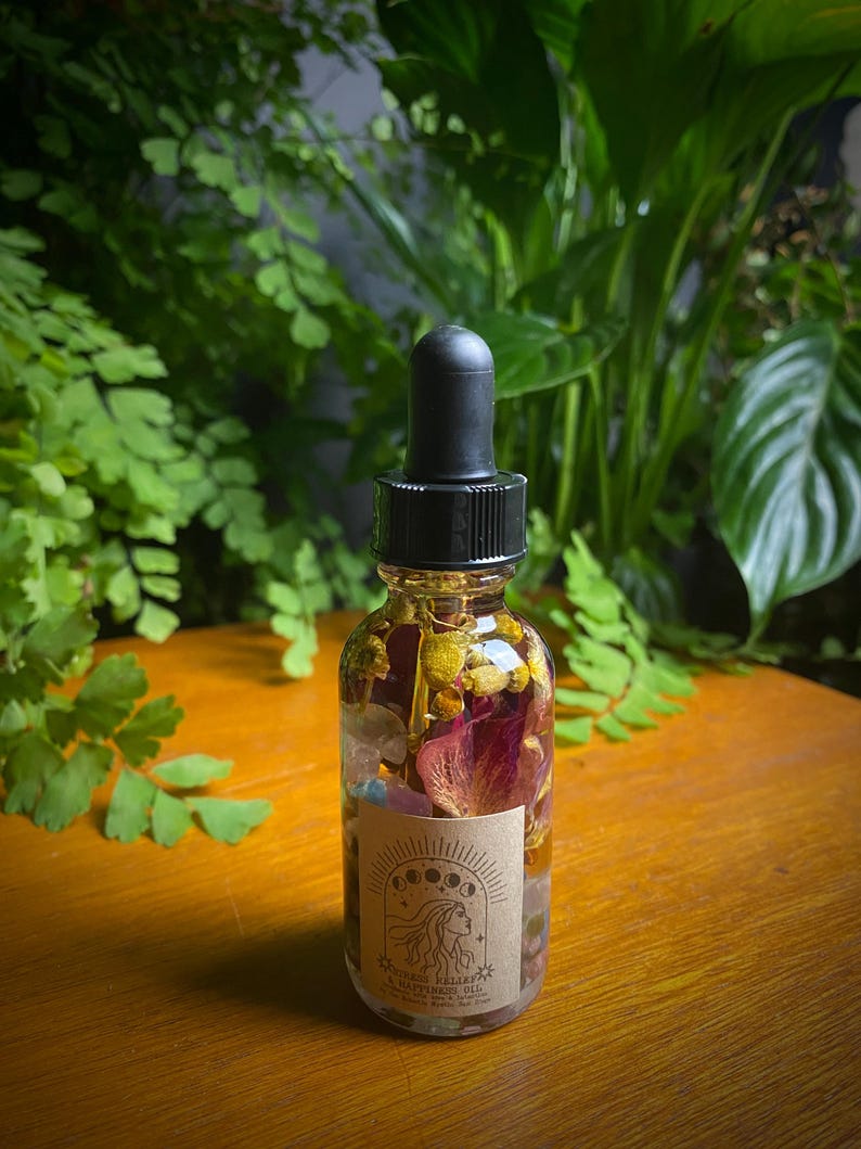 May include: A clear glass bottle with a black dropper top filled with a golden liquid and dried flowers and crystals. The label reads "WITCHY DAILY HAPPINESS OIL".