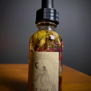 May include: A clear glass bottle filled with oil and dried flowers, topped with a black dropper. The label reads "SELF LOVE OIL" with an illustration of a woman under a crescent moon. The oil is a golden color, and the flowers are yellow, pink, and purple.