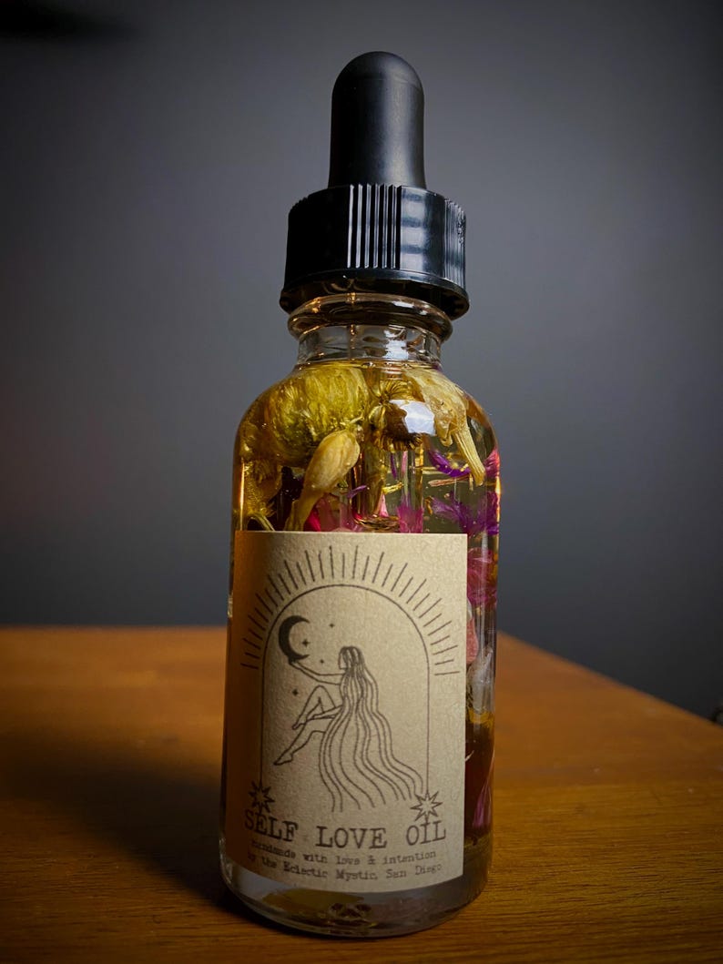 May include: A clear glass bottle filled with oil and dried flowers, topped with a black dropper. The label reads "SELF LOVE OIL" with an illustration of a woman under a crescent moon. The oil is a golden color, and the flowers are yellow, pink, and purple.