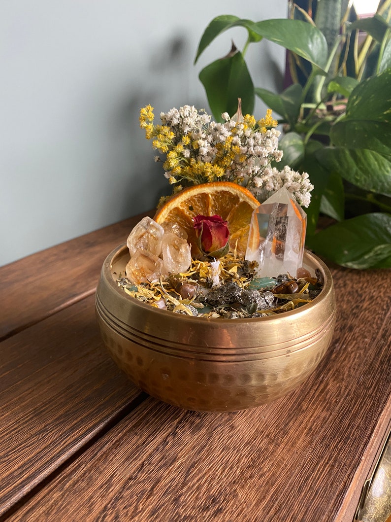 May include: A brass bowl filled with dried flowers, crystals, and an orange slice. The bowl has a hammered texture and is sitting on a wooden surface.
