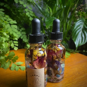 May include: Two small glass bottles filled with dried flowers and crystals. The bottles have black dropper tops and labels that say "Crystal Valley Happiness Oil".