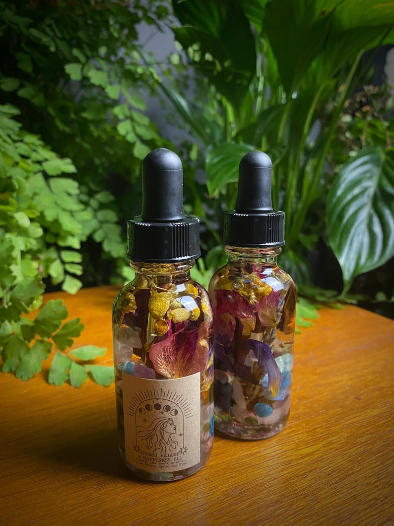 May include: Two small glass bottles filled with dried flowers and crystals. The bottles have black dropper tops and labels that say "Crystal Valley Happiness Oil".