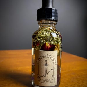 May include: A clear glass bottle filled with oil and dried herbs, topped with a black dropper. A tan label features a sword design and the words "CORD CUTTING OIL". The bottle is on a wooden surface.