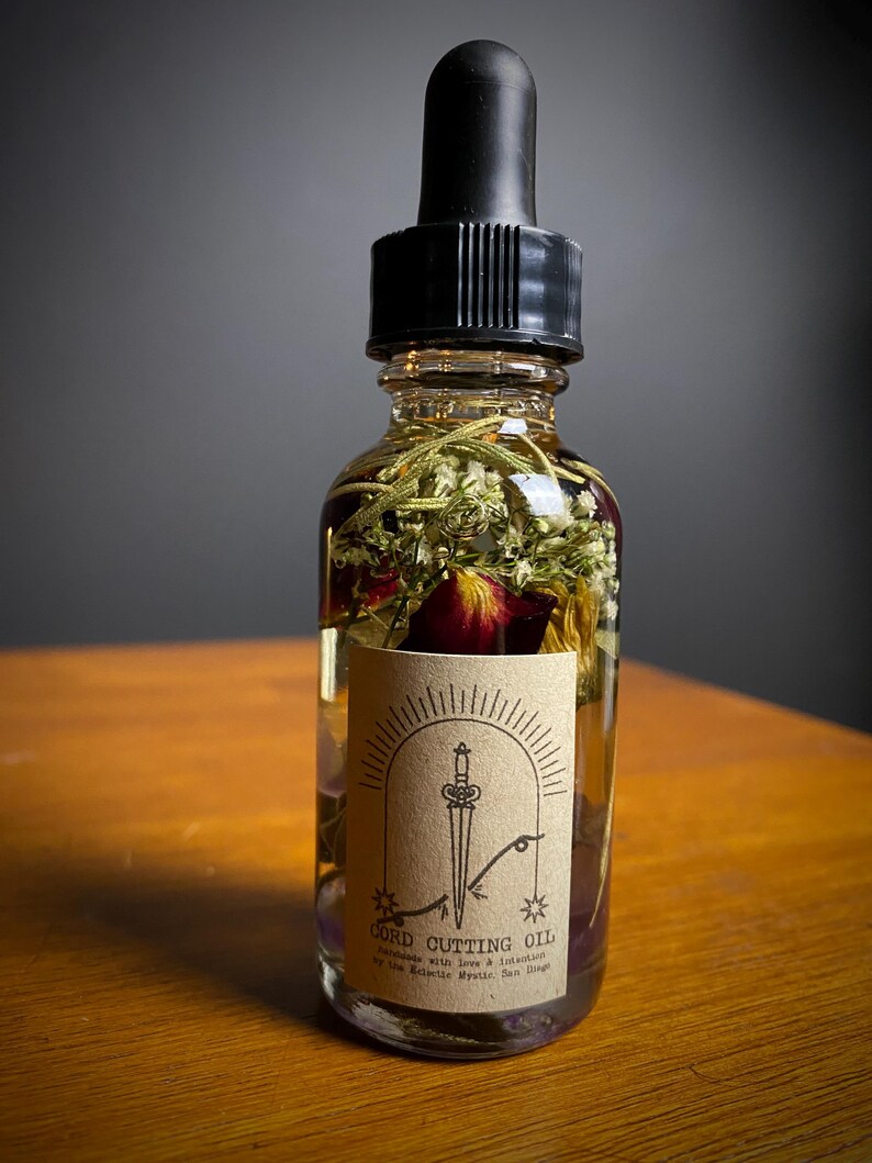 May include: A clear glass bottle filled with oil and dried herbs, topped with a black dropper. A tan label features a sword design and the words "CORD CUTTING OIL". The bottle is on a wooden surface.