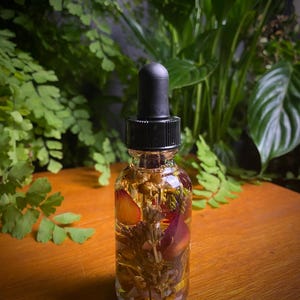 May include: A clear glass bottle with a black dropper top filled with a light yellow liquid and dried flowers and herbs. The bottle is sitting on a wooden surface.