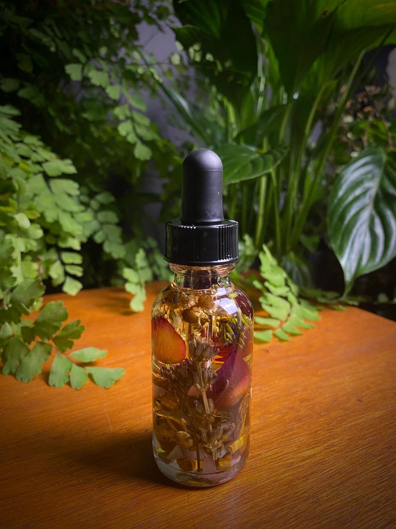 May include: A clear glass bottle with a black dropper top filled with a light yellow liquid and dried flowers and herbs. The bottle is sitting on a wooden surface.