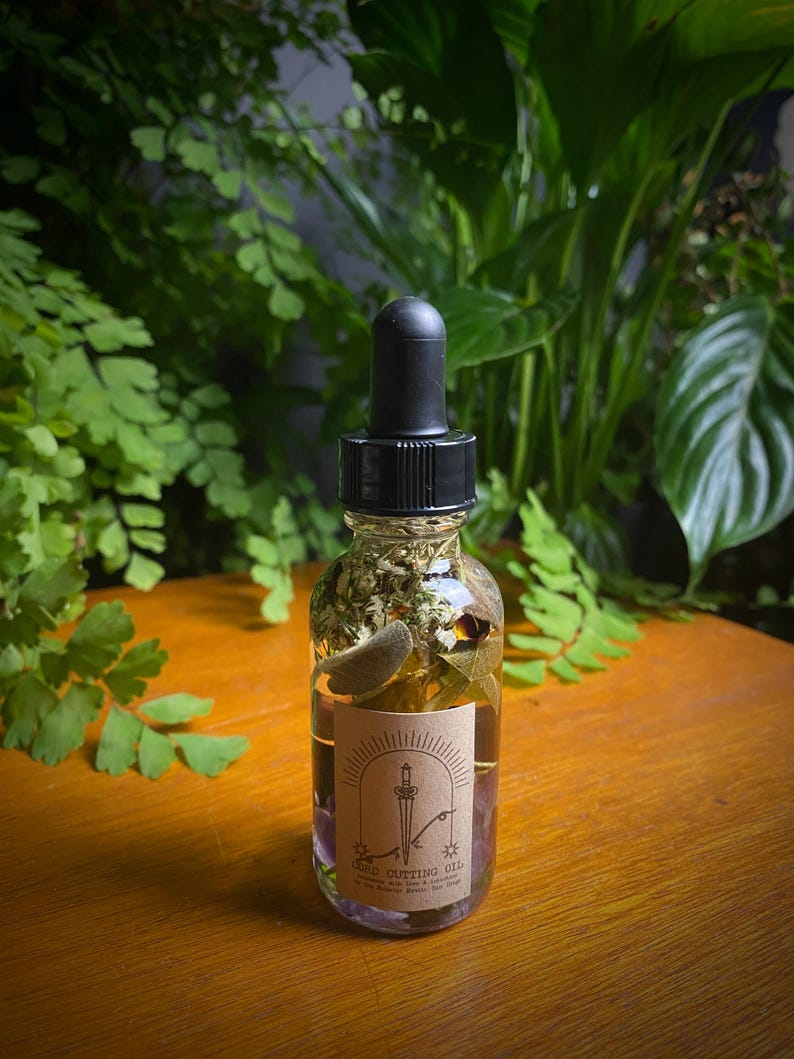 May include: A clear glass bottle with a black dropper top, filled with liquid and herbs. A label on the bottle reads "Cord Cutting Oil" with a sword and sun design. The bottle is on a wooden surface, with green foliage in the background.