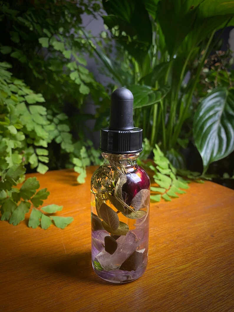 May include: A clear glass bottle with a black dropper top, filled with a clear liquid and botanical elements. The bottle contains purple crystals, green leaves, and a dark red flower. The background features green foliage.
