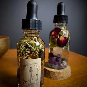 May include: Two glass bottles filled with oil and botanicals. One bottle has a label that reads "Cord Cutting Oil" with a sword illustration. The bottles have black droppers and are filled with herbs, flowers, and crystals. The bottles sit on a wooden surface.