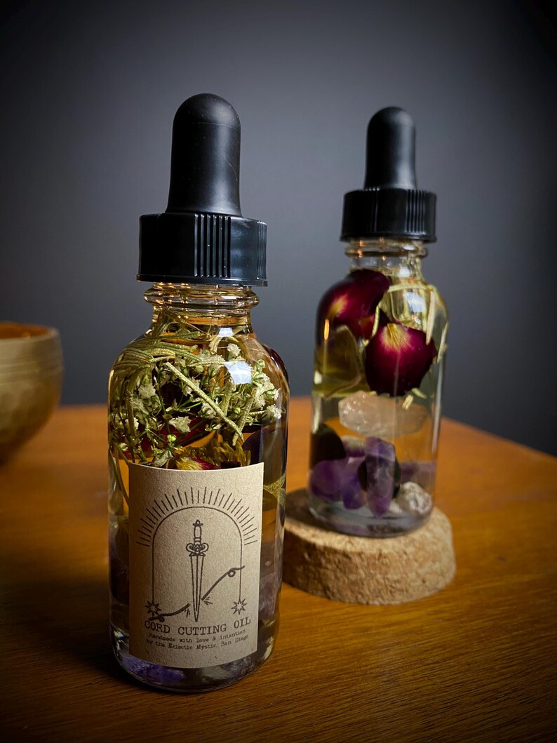 May include: Two glass bottles filled with oil and botanicals. One bottle has a label that reads "Cord Cutting Oil" with a sword illustration. The bottles have black droppers and are filled with herbs, flowers, and crystals. The bottles sit on a wooden surface.