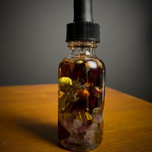 May include: A clear glass dropper bottle filled with a golden-brown liquid and various botanicals, including flowers and crystals. The bottle has a black dropper top and sits on a wooden surface.