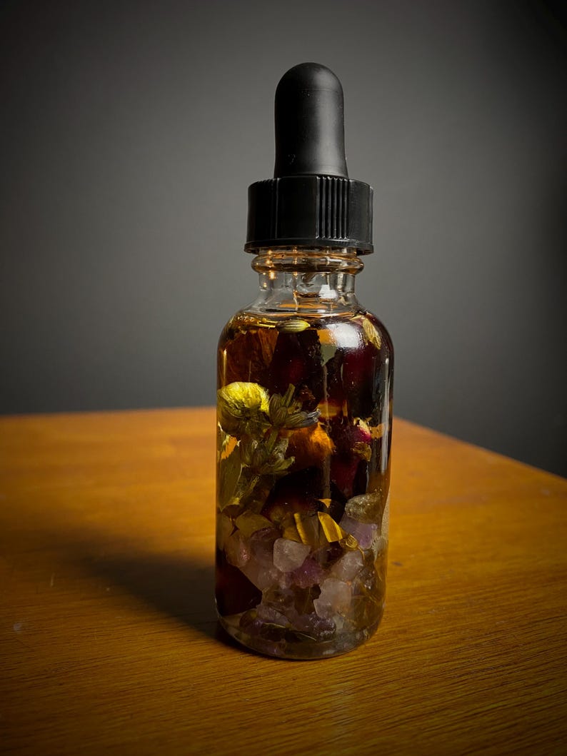 May include: A clear glass dropper bottle filled with a golden-brown liquid and various botanicals, including flowers and crystals. The bottle has a black dropper top and sits on a wooden surface.