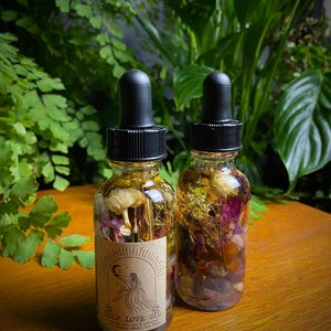 May include: Two glass dropper bottles filled with a clear liquid and botanical elements. The bottles have black caps and labels that read "SELF LOVE OIL" with an illustration of a woman. The background features lush green plants.