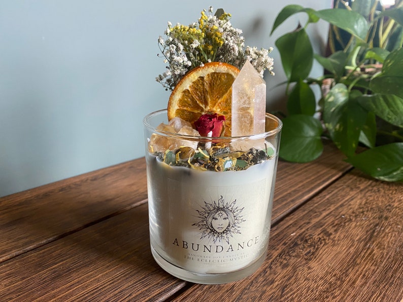 May include: A white candle in a glass jar with a dried orange slice, flowers, and crystals on top. The candle is labeled "Abundance Handmade Soy Candles The Eclectic Mystic".