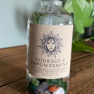 Puede incluir: Una botella de vidrio transparente con una etiqueta marr&oacute;n que dice "COURAGE & EMPOWERMENT" y "CRYSTAL INFUSED & FULL MOON CHARGED 4 OZ REFILLABLE MISTING SPRAY CREATED BY THE ECLECTIC MYSTIC SAN DIEGO, CA". La botella est&aacute; llena de cristales de colores.