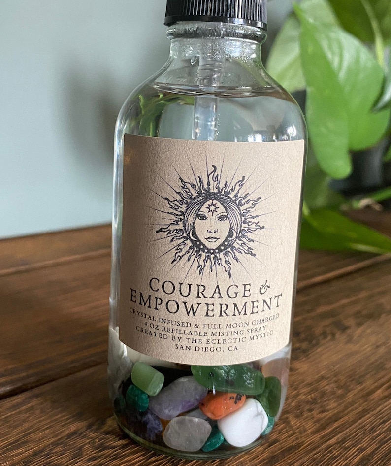 Puede incluir: Una botella de vidrio transparente con una etiqueta marr&oacute;n que dice "COURAGE & EMPOWERMENT" y "CRYSTAL INFUSED & FULL MOON CHARGED 4 OZ REFILLABLE MISTING SPRAY CREATED BY THE ECLECTIC MYSTIC SAN DIEGO, CA". La botella est&aacute; llena de cristales de colores.