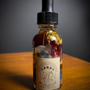 May include: A glass bottle with a dropper top filled with a clear liquid and various dried flowers and crystals. The bottle has a label that reads "Stress Relief & Happiness Oil" with a moon and star design.