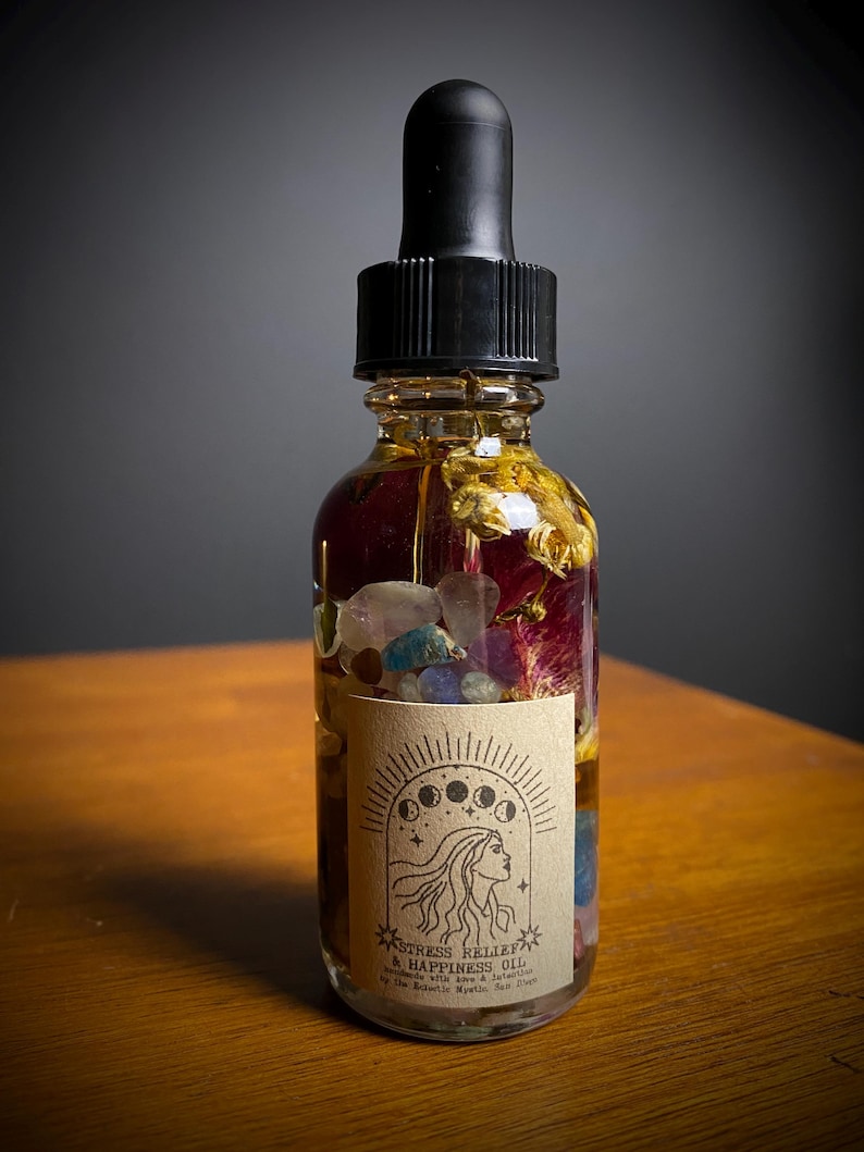 May include: A glass bottle with a dropper top filled with a clear liquid and various dried flowers and crystals. The bottle has a label that reads "Stress Relief & Happiness Oil" with a moon and star design.