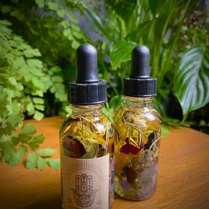 May include: Two small glass bottles filled with dried flowers and crystals. The bottles have black dropper tops and are labeled "Protection Oil".