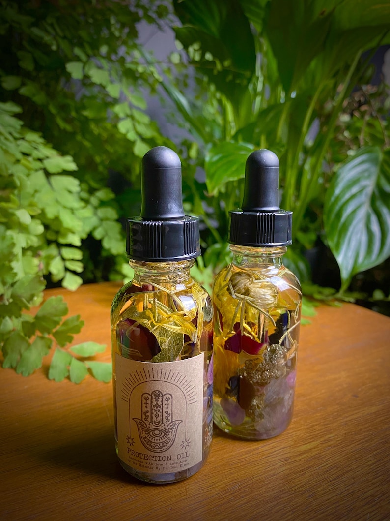 May include: Two small glass bottles filled with dried flowers and crystals. The bottles have black dropper tops and are labeled "Protection Oil".