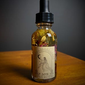 May include: A clear glass bottle with a black dropper top filled with a yellow liquid and dried flowers. The bottle has a brown label with the text "SELF LOVE OIL" and a drawing of a woman sitting with her legs crossed, holding a crescent moon.
