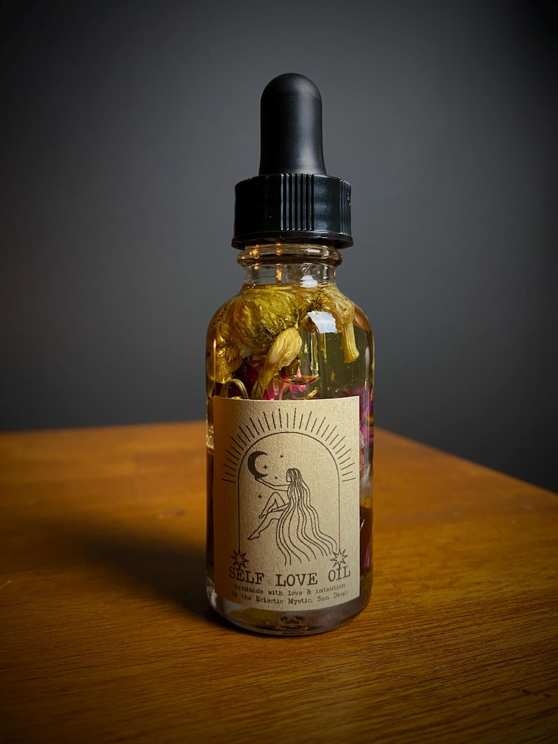 May include: A clear glass bottle with a black dropper top filled with a yellow liquid and dried flowers. The bottle has a brown label with the text "SELF LOVE OIL" and a drawing of a woman sitting with her legs crossed, holding a crescent moon.