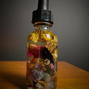 May include: A clear glass bottle with a black dropper top filled with a golden liquid and various crystals, including amethyst, rose quartz, and citrine.
