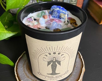 May include: Black candle with a variety of crystals embedded in the wax. The candle is on a brown agate coaster. The label on the candle reads "Strength, Courage & Confidence".