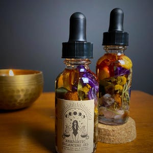 May include: Two small glass bottles filled with dried flowers, herbs, and crystals. The bottles have black dropper tops and brown labels that read "Energetic Balancing Oil" with a stylized image of a woman holding a scale.