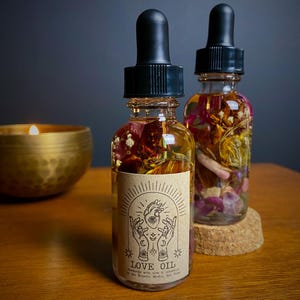 May include: Two small glass bottles with dropper tops filled with a clear liquid and dried flowers. The bottles have brown paper labels with a black and white illustration of a hand holding a heart with an eye in the centre. The label on the front bottle reads "LOVE OIL Handmade with love & intention by The Eclectic Mystic, New Orleans."