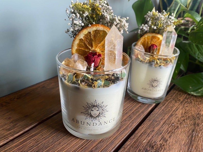 May include: Two glass candle jars with white candles, topped with dried orange slices, white flowers, and clear quartz crystals. The jars have a black and white label that reads "Abundance".