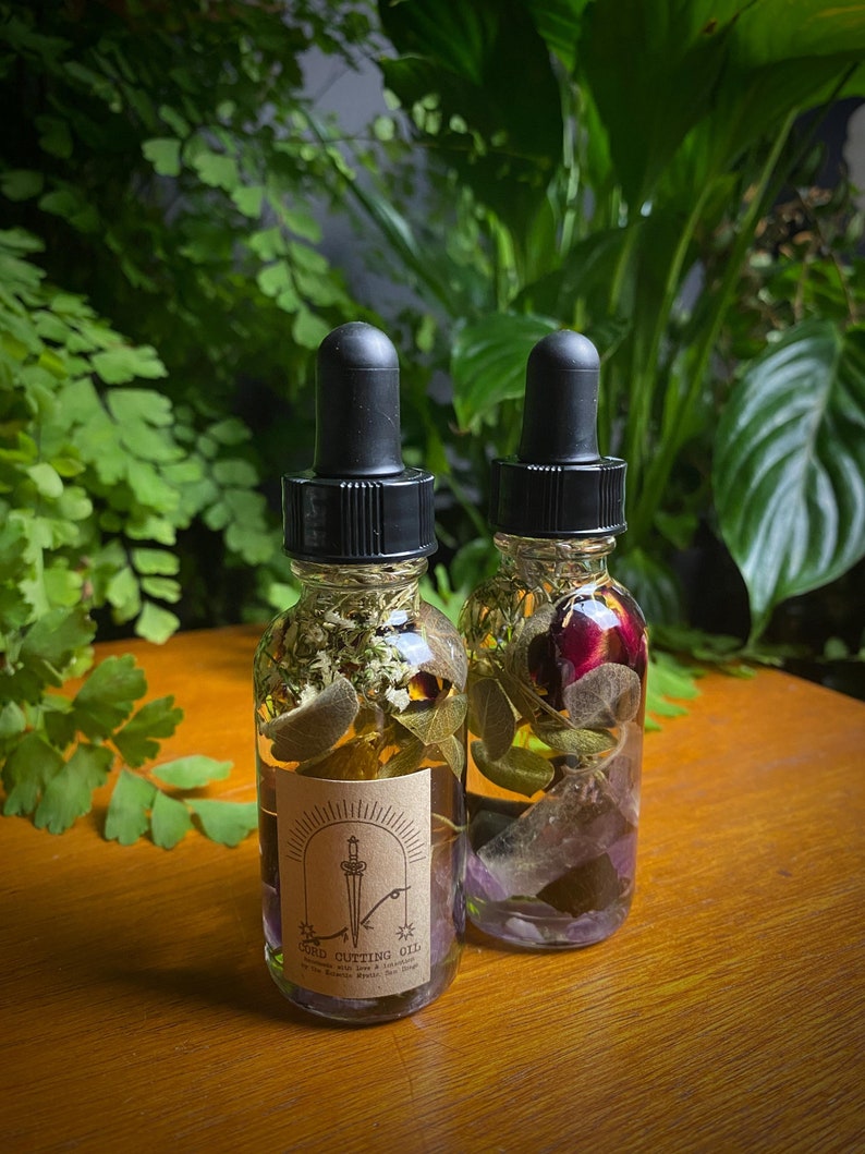 May include: Two glass bottles of Cord Cutting Oil with black droppers. The bottles are filled with herbs and flowers. One bottle has a label with a sword design. The background features green plants and a wooden surface.