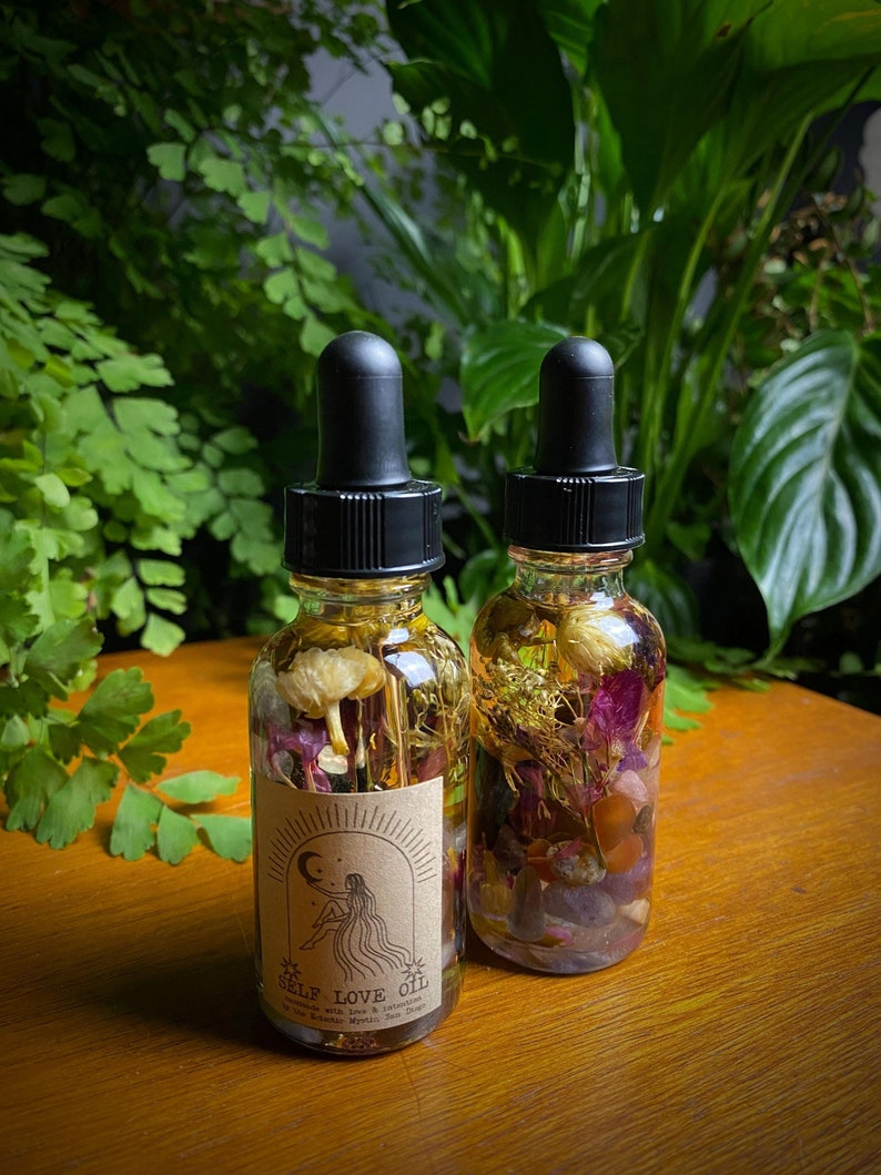 May include: Two small glass bottles filled with dried flowers, herbs, and crystals. The bottles have black dropper tops and brown labels that say "Self Love Oil" with a moon and star design.