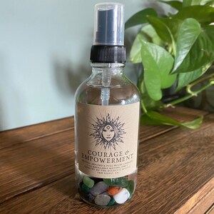 Puede incluir: Una botella de vidrio transparente con un pulverizador negro llena de un l&iacute;quido transparente y cristales. La botella tiene una etiqueta marr&oacute;n con el texto "COURAGE & EMPOWERMENT" y un sol con un dise&ntilde;o de cara.